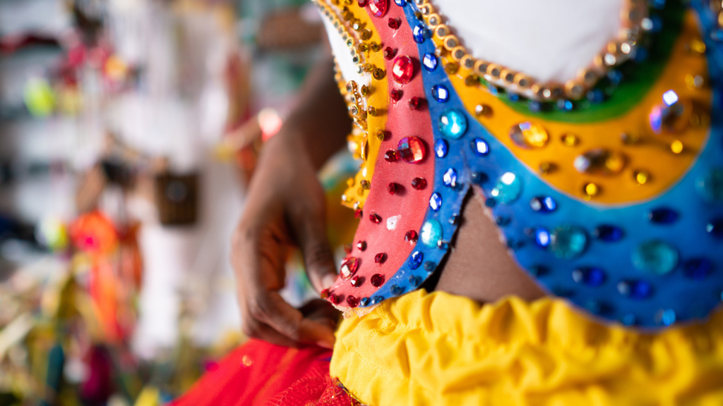 Close-up detalhado de uma fantasia de Carnaval sendo ajustada. A peça é vibrante, com cores do arco-íris, pedrarias brilhantes e lantejoulas aplicadas. A imagem ilustra o conceito de personalização e tutoriais, uma tática eficaz de marketing de carnaval para estimular o engajamento do público.
