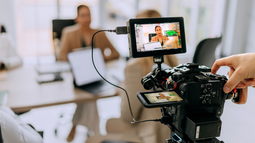 Visão por trás de uma câmera de vídeo profissional em um tripé, filmando uma mulher sentada em um escritório moderno. A tela de monitoramento da câmera mostra o enquadramento da apresentadora, enquanto ao fundo, fora de foco, vê-se o ambiente de trabalho com laptops. Uma mão ajusta as configurações da câmera, simbolizando a produção de conteúdo direcionado.