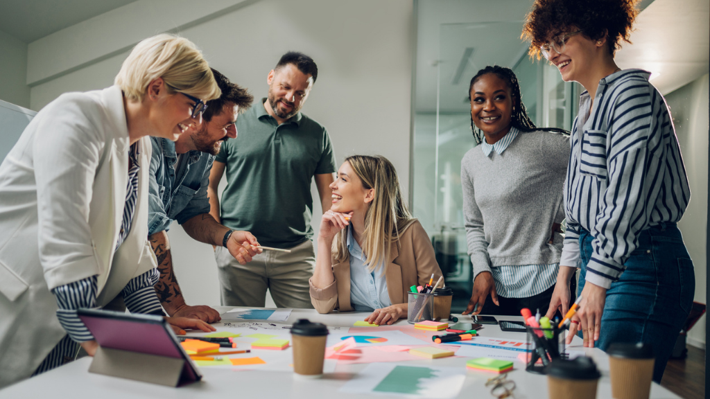 Grupo de profissionais sorridentes em uma reunião de brainstorming ao redor de uma mesa branca. A mesa está repleta de post-its coloridos, gráficos impressos, canetas e café, simbolizando a fase de ideação e troca de insights para uma estratégia de marketing de carnaval.