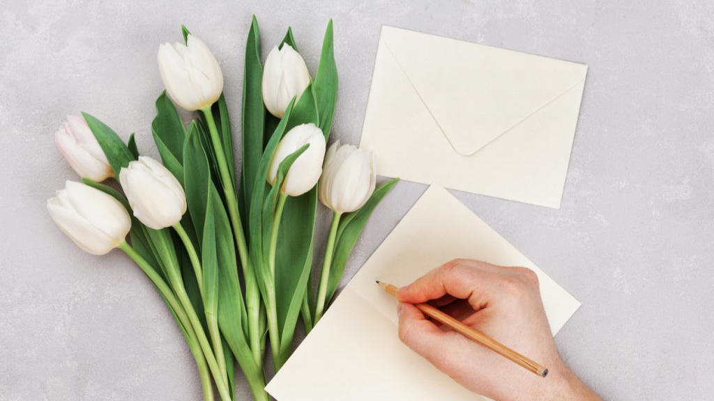 Vista de cima de uma mão masculina escrevendo com lápis em um cartão em branco, ao lado de um envelope bege e um buquê de tulipas brancas frescas sobre um fundo cinza texturizado. A imagem ilustra o conceito de humanização e personalização em estratégias de marketing para 2026.