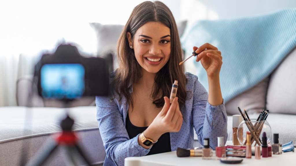Uma jovem sorridente com cabelos castanhos grava um vídeo de maquiagem. Ela segura um aplicador de corretivo em frente a uma câmera profissional montada em um tripé. Sobre a mesa branca à sua frente, há diversos cosméticos como batons, pincéis e esmaltes. O ambiente é uma sala de estar iluminada e aconchegante.