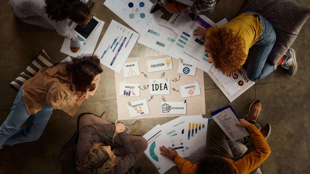 Vista aérea de uma equipe diversa reunida em círculo no chão, colaborando em um painel de ideias centralizado com a palavra "IDEA". Ao redor, há diversos gráficos, fluxogramas e documentos de planejamento estratégico que auxiliam no desenvolvimento de uma campanha de marketing de carnaval.