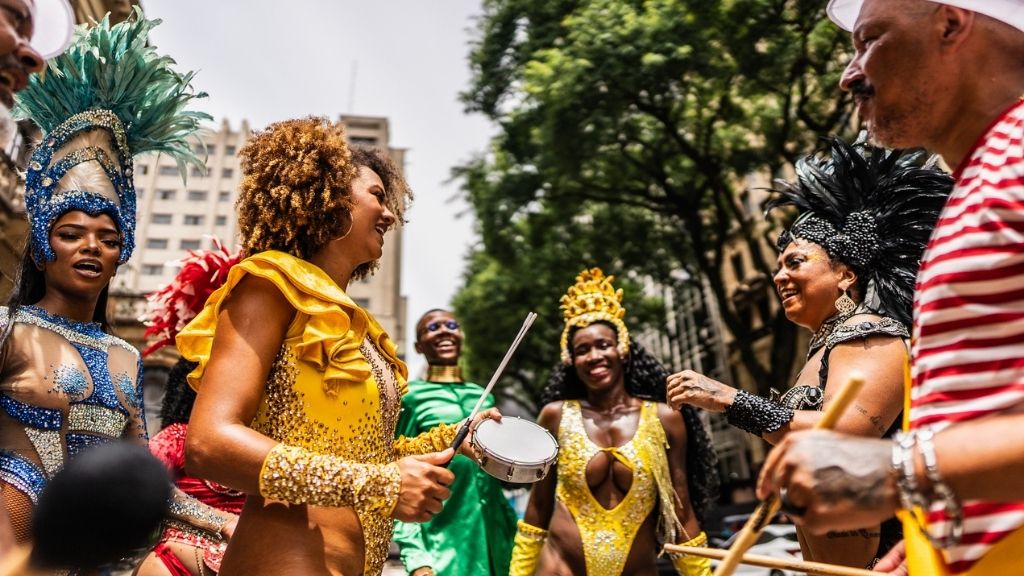 Fotografia vibrante e em close de um desfile de Carnaval de rua. Em destaque, um homem sorridente toca um pandeiro enquanto passistas com fantasias luxuosas de penas vermelhas e amarelas dançam ao fundo. A imagem captura a essência da festa e a alegria do público, ideal para ilustrar um artigo sobre marketing de carnaval.