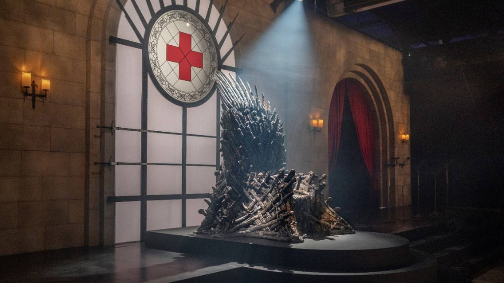 Réplica do Trono de Ferro da série Game of Thrones posicionado em um cenário que simula um castelo medieval. Ao fundo, um vitral circular exibe uma cruz vermelha, símbolo de doação de sangue, em vez de um brasão de família. A iluminação é dramática, com um feixe de luz focado no trono. A imagem ilustra a campanha da HBO "Brindes em troca de propósito", unindo entretenimento e responsabilidade social como estratégia de marketing para 2026.