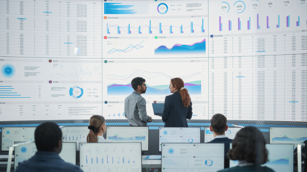 Vista traseira de uma equipe de analistas sentados em workstations em um escritório moderno, de frente para uma imensa parede de telas curvas que exibe múltiplos dashboards de dados, gráficos coloridos de barras e linhas, e tabelas financeiras detalhadas. No centro, dois profissionais de pé discutem as análises, ilustrando a escala massiva de dados e o monitoramento de tráfego inválido realizado por empresas de conformidade digital.