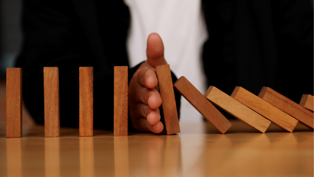 Close em uma mão humana de uma pessoa vestindo um terno preto, posicionada verticalmente no meio de uma fileira de blocos de madeira sobre uma mesa clara. A mão atua como uma barreira, impedindo que os blocos que já caíram à direita atinjam os blocos que ainda estão de pé à esquerda. A imagem é uma metáfora para a interrupção do prejuízo e a proteção contra o tráfego inválido em estratégias de marketing digital.