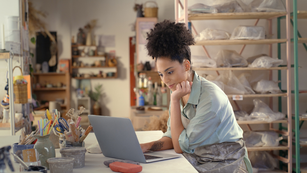 Uma artesã de cerâmica negra sorridente, com cabelo afro, sentada em seu ateliê cheio de prateleiras e ferramentas, olhando para a tela de um notebook onde ela está usando a plataforma Pomelli para transformar rapidamente uma foto simples de suas peças em uma imagem de estúdio profissional para marketing.