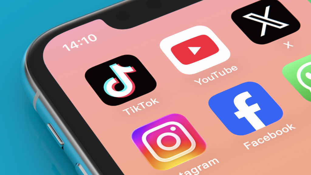 Close-up em ângulo fechado da tela de um smartphone moderno, exibindo uma grade de ícones coloridos de aplicativos populares sobre um fundo escuro desfocado com luzes neon rosa. No topo, os ícones azuis do Facebook e o gradiente rosa e roxo do Instagram. No meio, o ícone preto e branco do X (antigo Twitter) flanqueado pelo verde do WhatsApp e o azul do Discord. Na parte inferior, o ícone laranja do Reddit, o vermelho do YouTube e o preto e branco do TikTok. A composição representa a interconexão do ecossistema digital e a presença oculta de tráfego inválido em plataformas de alto engajamento.