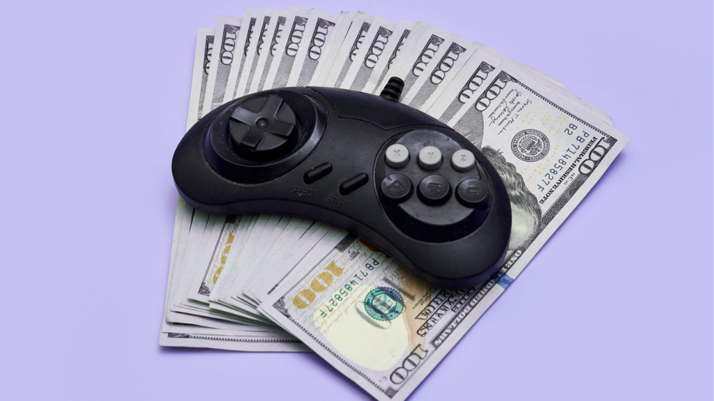 Uma foto em ângulo fechado com fundo lilás claro sólido. Um controle de videogame retrô preto, com um direcional e quatro botões principais (A, B, C, Y), está posicionado diretamente sobre um maço espalhado de notas de 100 dólares americanos autênticas. O controle parece "pesar" sobre o dinheiro. A imagem simboliza como o setor de Gaming e os aplicativos MFA são alvos para a drenagem de orçamentos publicitários através de tráfego inválido.