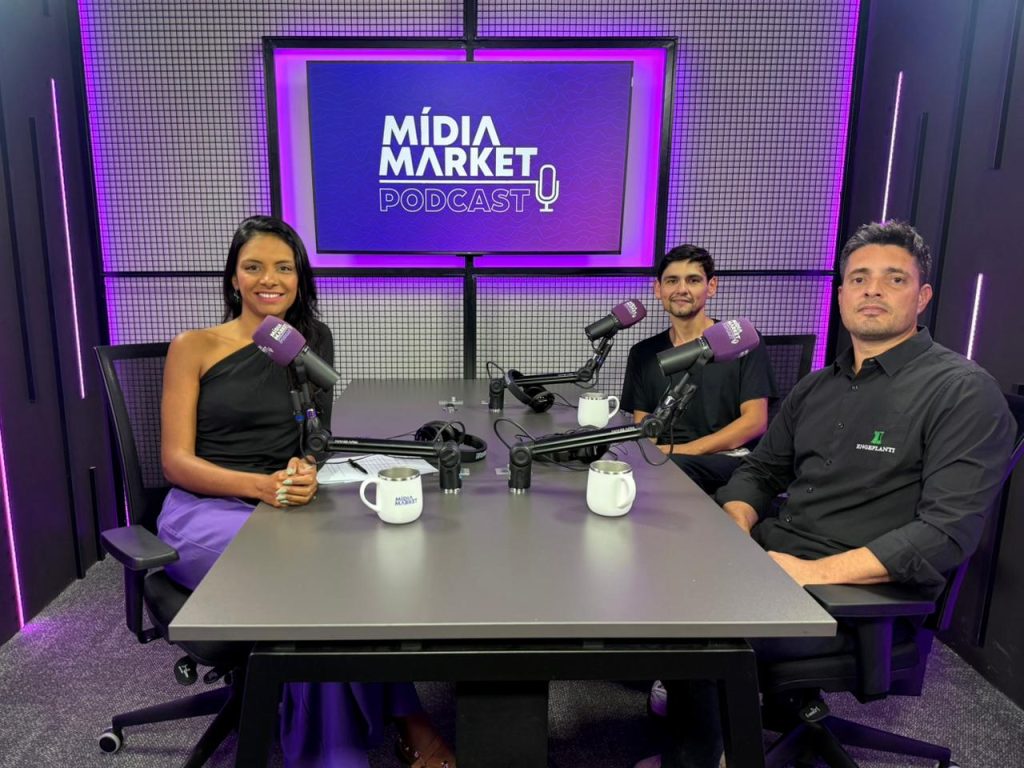 Três pessoas sentadas a mesa para entrevista de podcast com cores em roxo sendo destacadas com uma mesa ao centro com canecas e microfones.