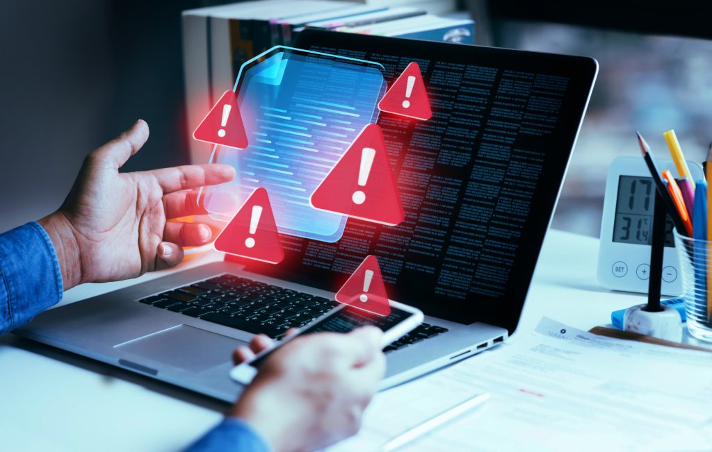 Foto em ângulo fechado de uma pessoa utilizando um notebook e um smartphone em uma mesa de escritório. Sobre a tela do computador, flutuam hologramas de ícones de alerta vermelhos com pontos de exclamação e a representação de um documento digital azul. A imagem simboliza erros em relatórios, avisos de fraude e a presença de tráfego inválido infectando dados digitais.