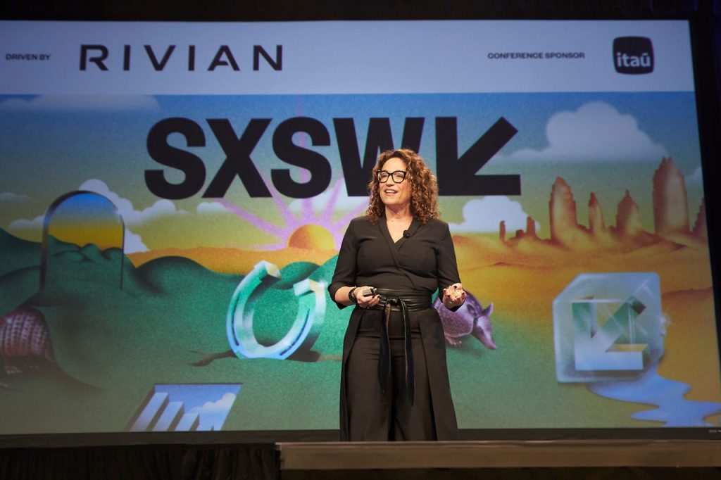 Fotografia em plano médio da futurista Amy Webb, usando óculos e roupa preta, palestrando no SXSW 2026. Ao fundo, um telão colorido exibe o logo do SXSW, montanhas estilizadas, um tatu e as marcas Rivian e Itaú. Ela segura um passador de slides e sorri.