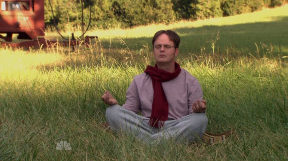 Fotografia de uma cena da série The Office com o personagem Dwight Schrute sentado de pernas cruzadas em posição de meditação no meio de um gramado verde. Ele está de olhos fechados, usa óculos, uma blusa cinza de manga longa e um cachecol vermelho. Ao fundo, árvores e uma carroça antiga de fazenda. Logo da emissora NBC discretamente no canto inferior esquerdo.