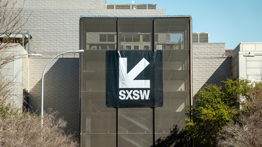 Fotografia externa em plano médio da fachada de um edifício moderno de concreto durante o dia. No centro, há uma grande lona preta estendida verticalmente com o logotipo branco do SXSW e sua seta característica apontando para baixo e para a esquerda. Árvores com poucas folhas emolduram as laterais inferiores da imagem sob um céu azul claro.