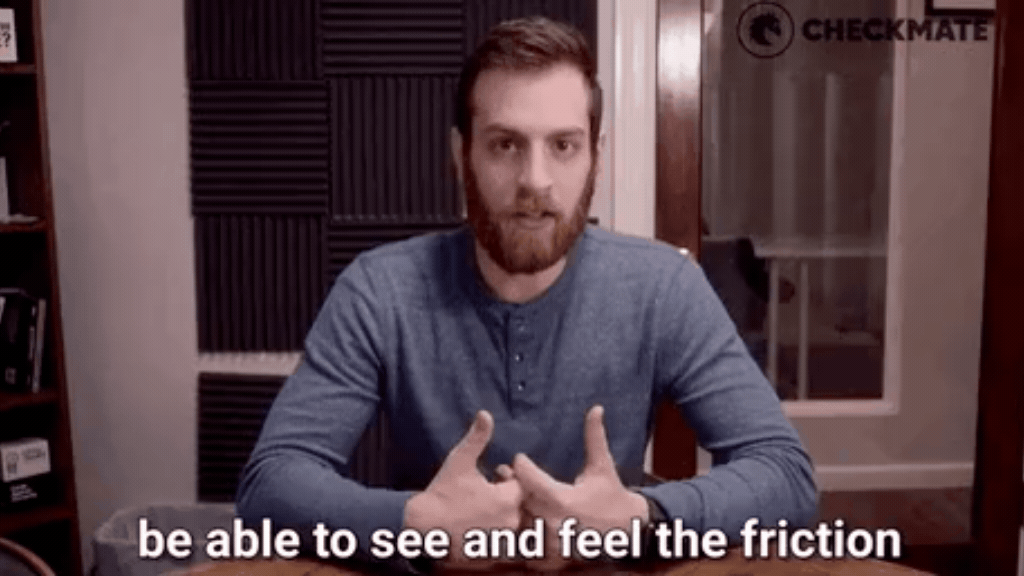 GIF de um homem jovem com barba e blusa de manga longa azul, sentado em frente a uma mesa de madeira em uma sala de reuniões. Ele gesticula com as mãos unidas e fala de forma séria. Na parte inferior, há uma legenda em letras brancas que diz: "be able to see and feel the friction" (ser capaz de ver e sentir a fricção).