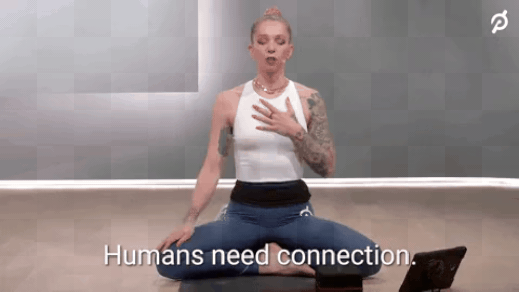 GIF de uma mulher loira com cabelos presos e braços tatuados, sentada de pernas cruzadas em um estúdio fazendo um exercício de respiração com a mão direita no peito. Na parte inferior, a legenda em letras brancas diz: "Humans need connection" (Humanos precisam de conexão). Logo da Peloton visível no canto superior direito.