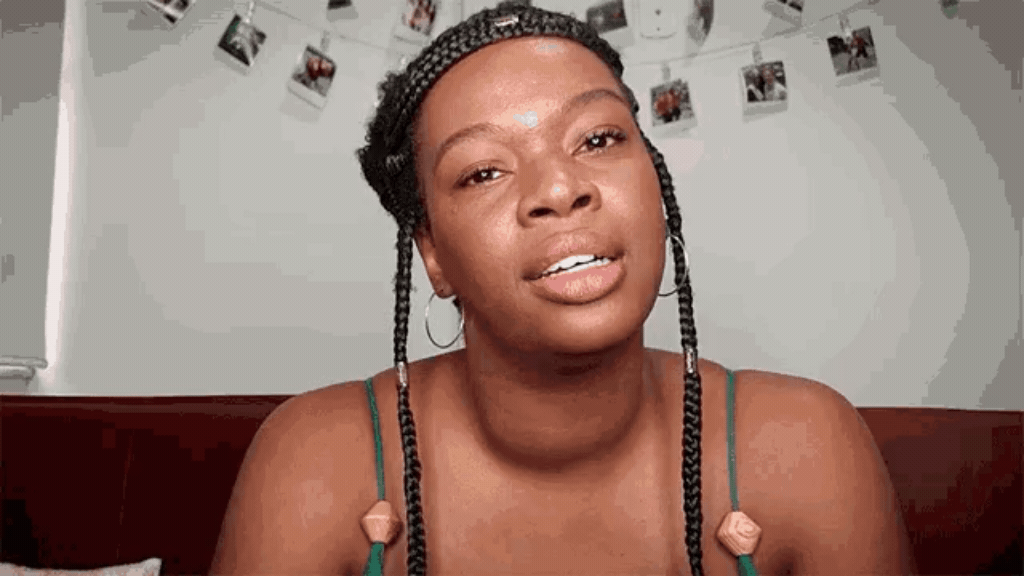 Mulher negra de tranças conversando para a câmera em um ambiente aconchegante com fotos penduradas ao fundo, representando a criação de conteúdo e a busca social entre diferentes gerações.