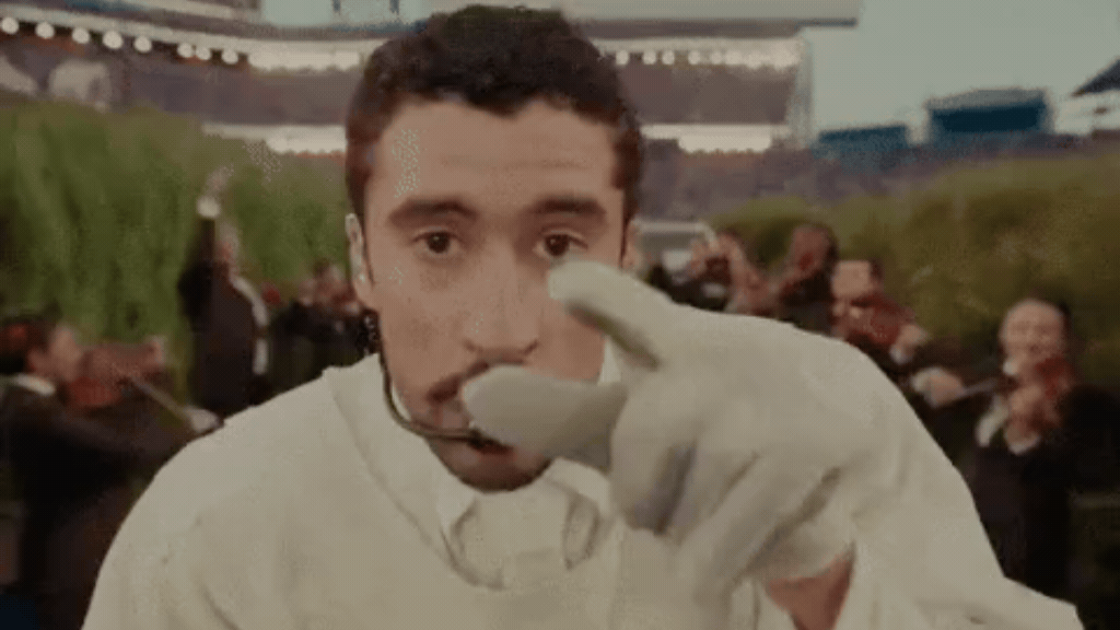 Uma captura de tela em plano médio de Bad Bunny olhando diretamente para a câmera e apontando o dedo indicador em luva branca para frente. Ele veste uma roupa clara e possui um microfone auricular. Ao fundo, de forma desfocada, latina há músicos vestidos de terno tocando violino em um ambiente externo.