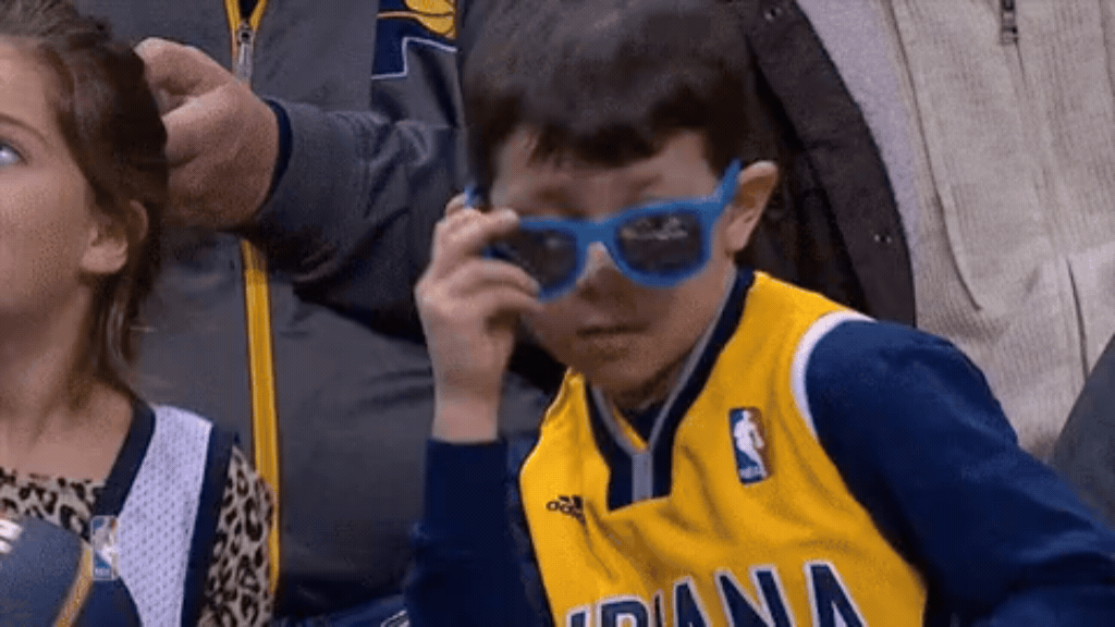 Uma foto de foco médio mostra um menino jovem sentado em uma arquibancada lotada de um evento esportivo. Ele tem cabelo curto e castanho e veste uma camisa de basquete azul e amarela do Indiana Pacers por cima de uma camisa de manga longa azul escura. Com a mão direita, ele ajusta ou abaixa levemente um par de óculos de sol azuis brilhantes, olhando diretamente para a câmera com uma expressão séria e confiante. À sua esquerda, parte do rosto e ombro de uma menina com estampa de leopardo é visível, e adultos desfocados estão atrás dele.