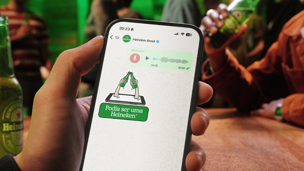 Mão segurando um smartphone que exibe uma conversa no WhatsApp com o chatbot da Heineken Brasil, mostrando um áudio encaminhado de mais de 4 minutos e uma figurinha com a frase 'Podia ser uma Heineken', com fundo de pessoas em um bar.