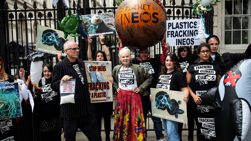A estilista e ativista Vivienne Westwood em protesto ambiental ao lado de ativistas segurando cartazes contra a poluição por plástico e fraturamento hidráulico, ilustrando a moda como manifesto político e expressão em tempos de saturação de mídia.