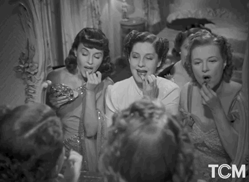 GIF em preto e branco de cinema clássico mostrando três mulheres em frente a um espelho retoque de maquiagem aplicando batom, ilustrando o conceito de Lipstick Effect e a saturação de mídia.